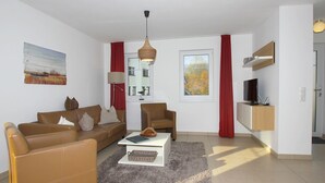 En moderne stue med sofa, lænestol og sofabord, et fjernsyn på en træhylde, og et vindue med udsigt til træer.