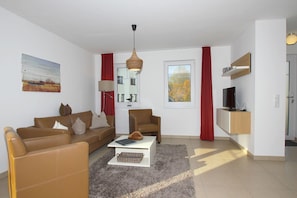 A modern living room with a sofa, armchair, and coffee table, a TV on a wooden shelf, and a window with a view of trees.