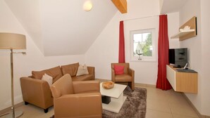 A modern living room with a brown leather sofa, a white coffee table, a red chair, and a TV on a wooden stand. - Beachfront vacation rental with a balcony, access to the pool and sauna - Rex Rugia - Rex Rugia (Göhren)