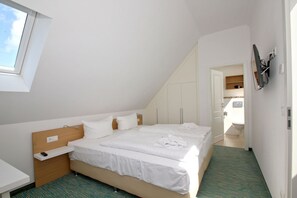 Ein modernes, helles Schlafzimmer mit einem großen Bett, einem Holzkopfteil und einem kleinen Nachttisch. Ein Fenster sorgt für Tageslicht und bietet Blick zum Himmel.