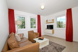A modern living room with a brown leather sofa, a white coffee table, and a TV mounted on the wall. Red curtains frame the windows, and a lamp is on the side table.