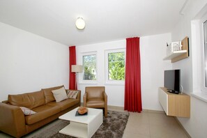 A modern living room with a brown leather sofa, a white coffee table, a leather armchair, a TV on a wooden stand, and red curtains. - Beachfront vacation rental with a terrace, access to the pool and sauna - Rex Rugia H-1010 - (Göhren)