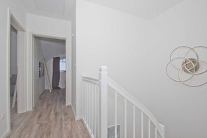 A hallway with a wooden floor, white walls, and a white railing. There is a door leading to another room and a wall-mounted circular object. - At the sailing marina, with a patio, garden, and ocean view—Boddentraum Abendrot vacation home— (Gager)