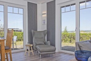 A modern room with a grey armchair, a small stool, and a glass table. Large windows offer a view of greenery and a building. - At the sailing marina, with a patio, garden, and ocean view—Boddentraum Abendrot vacation home— (Gager)