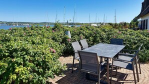An outdoor patio with a table and chairs, surrounded by lush greenery and overlooking a marina with sailboats. - At the sailing harbor, terrace, garden, sea view - vacation home Boddentraum Morgenrot - (Gager)
