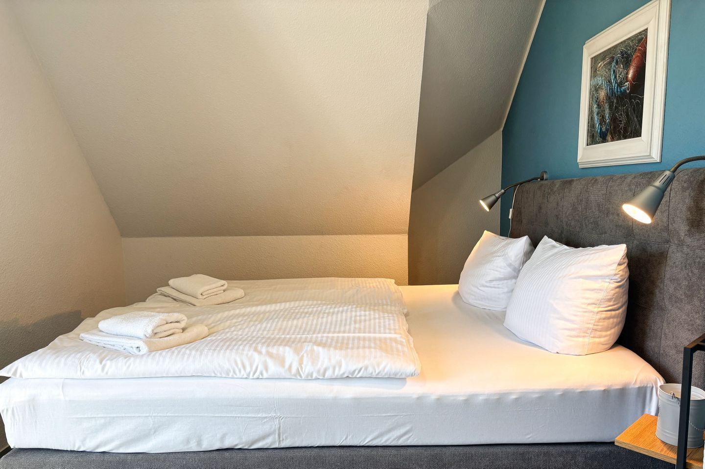 A neatly made bed with white linens and towels, a headboard lamp, and a painting on the wall.