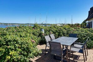 Un patio al aire libre con una mesa y sillas, rodeado de vegetación exuberante y vistas a un puerto deportivo con veleros.