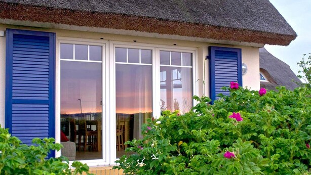 A house with a thatched roof, blue shutters, and a glass door leading to an interior with a view of the sunset. - At the sailing harbor, terrace, garden, sea view - vacation home Boddentraum Morgenrot - (Gager)