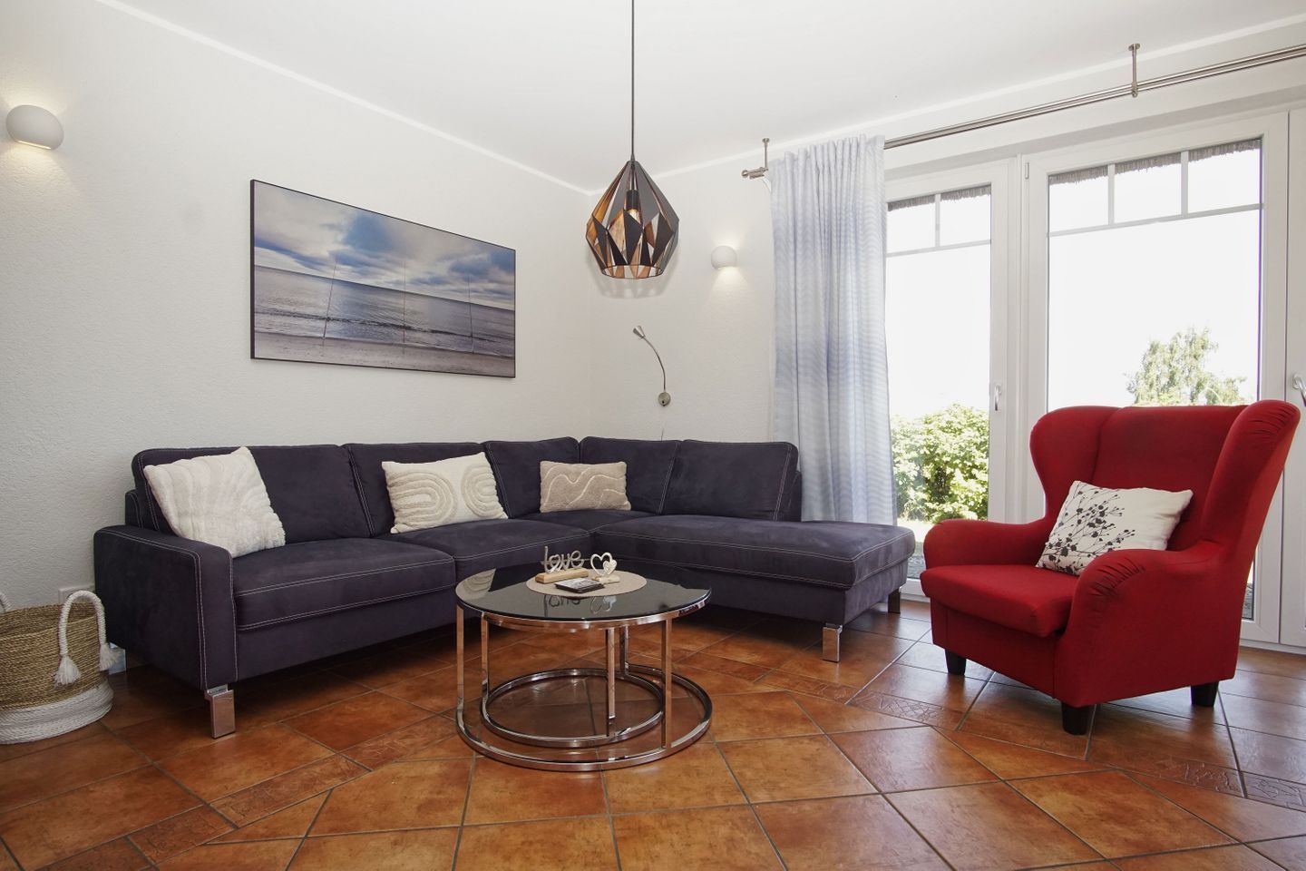 Ein Wohnzimmer mit einem dunklen Sofa, einem roten Sessel, einem runden Couchtisch, einem Bild an der Wand und einem großen Fenster mit Vorhängen.