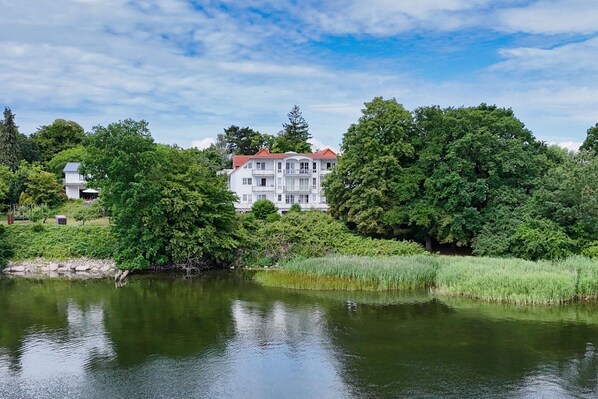 A large, multi-story building with a red roof, surrounded by lush greenery and a calm body of water. - 2-room vacation apartment with sea view from the balcony - Villa Vilmblick FeWo 15 - Villa (Putbus)