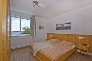 A bedroom with a wooden bed, a nightstand, a window with a view of the sea, and a painting on the wall. - 2-room vacation apartment with a view of the sea from the balcony - Villa Vilmblick FeWo 15 - Villa (Putbus)