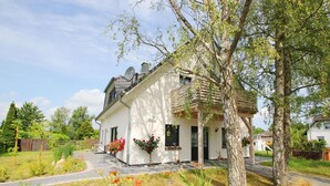 Une maison à deux étages, à l’extérieur blanc, dotée d’un porche et d’un jardin où poussent diverses plantes et fleurs.