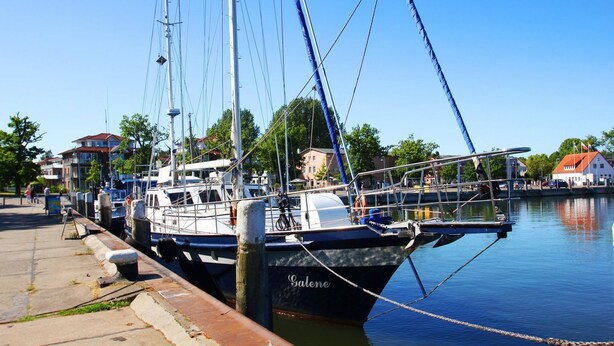 Un voilier, baptisé « Galene », est amarré dans une marina, avec des bâtiments et des arbres en arrière-plan.