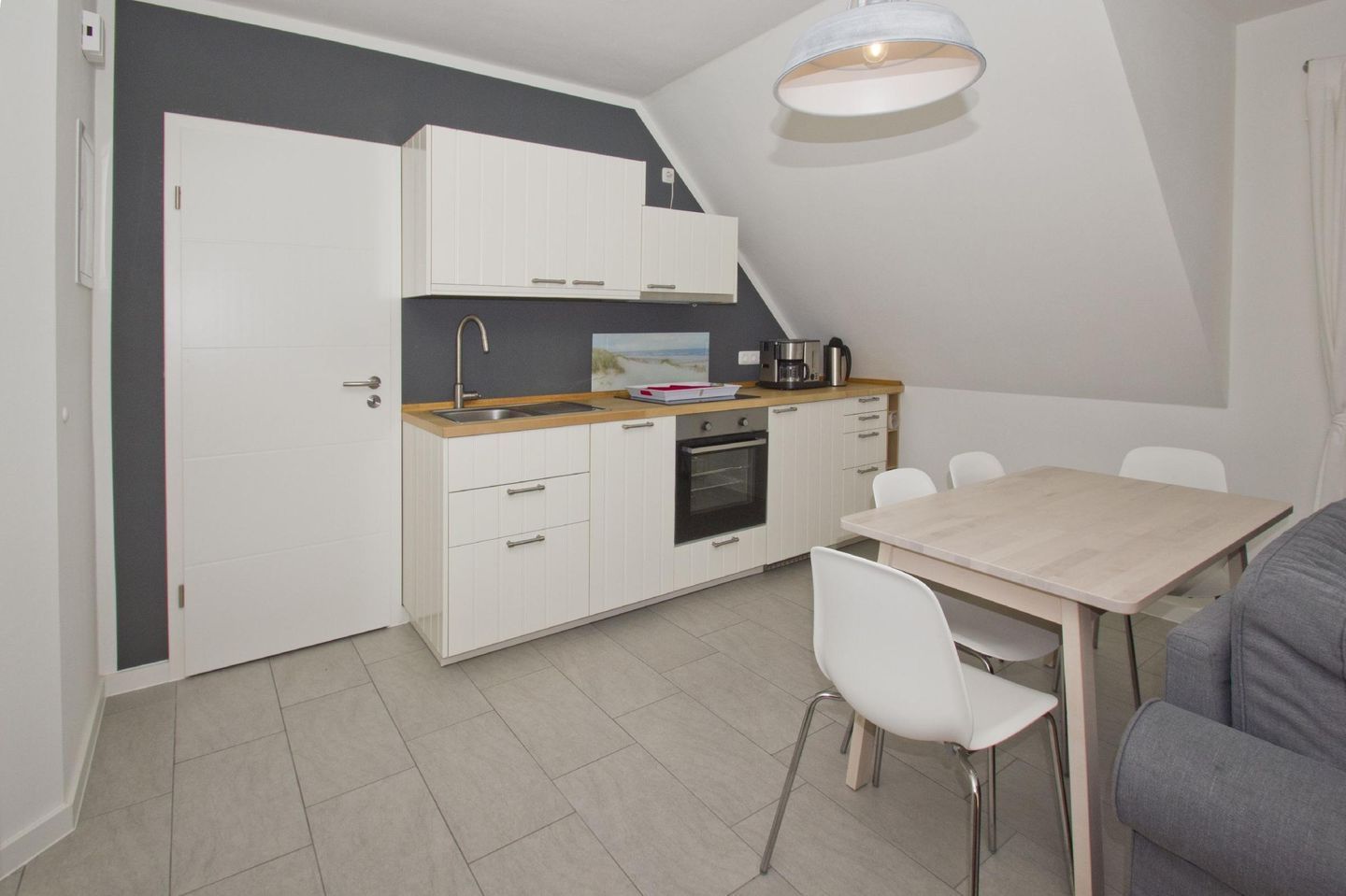 A compact kitchenette with white cabinets, a sink, and a small oven. There is a dining table with chairs and a light fixture above.