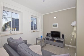 Ein modernes Wohnzimmer mit einem grauen Sofa, einem Fernseher und einem kleinen Tisch mit Lautsprechern.