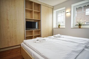 Un dormitorio con una cama, un televisor sobre una estantería de madera y una ventana con plantas.