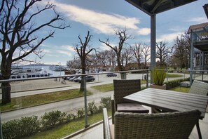 A covered outdoor seating area with wicker furniture, overlooking a road with parked cars and a boat docked at a marina. - Hafenresidenz Lauterbach Wohnung 2-3 - Hafenresidenz (Putbus)