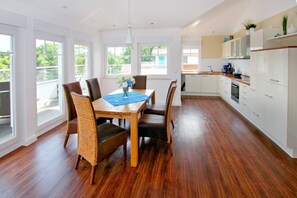 Una sala da pranzo spaziosa con tavolo e sedie in legno, ampie finestre e una cucina moderna con mobili bianchi.