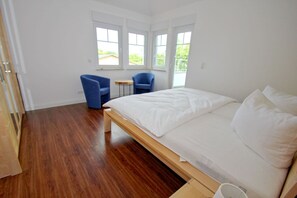 A bedroom with a wooden bed, two blue chairs, a small round table, and large windows with a view of greenery.