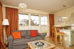 A living room with a grey sofa, orange cushions, a glass coffee table, and a kitchen area with a wooden chair and a white refrigerator. - Directly behind the dune, with balcony and sea view - Strandvilla Böck FeWo 10 - (Glowe)