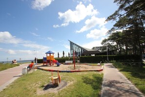 A playground with slides and swings, a paved path, and a building in the background. - Directly behind the dune, with balcony and sea view - Strandvilla Böck FeWo 10 - (Glowe)