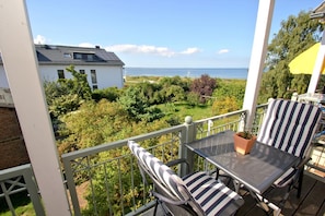 A balcony with a table and two striped chairs, overlooking a garden and a body of water. - Directly behind the dune, with balcony and sea view - Strandvilla Böck FeWo 13 - (Glowe)