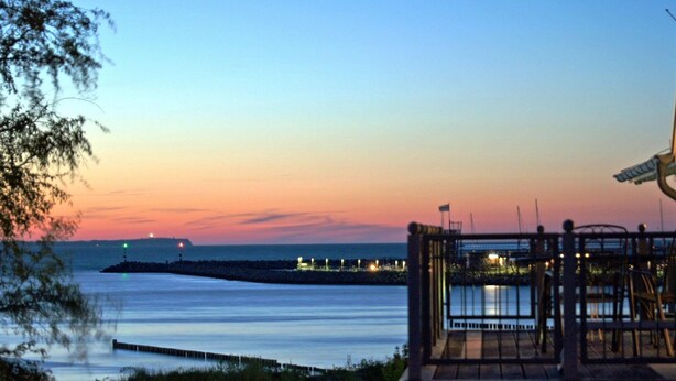 Une vue sur la côte, avec une jetée qui s’avance dans l’eau, un bâtiment doté d’un balcon donnant sur la mer, et un coucher de soleil en arrière-plan.
