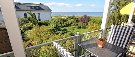 A balcony with a table and two striped chairs, overlooking a garden and a body of water.