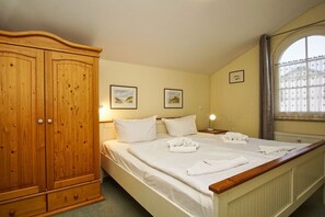 A bedroom with a large wooden wardrobe, a bed with neatly folded towels, two framed pictures on the wall, and a window with a curtain. - Directly behind the dune, with balcony and sea view - Strandvilla Böck FeWo 15 - (Glowe)