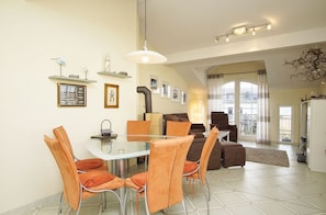 A dining area with a glass table and orange chairs, a sofa, and a chandelier.
