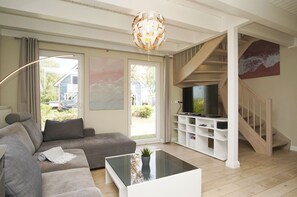 A living room with a grey sofa, a glass coffee table, a TV on a white entertainment unit, and a staircase with a wooden railing.