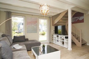 A living room with a grey sofa, a glass coffee table, a TV on a white entertainment unit, and a staircase with a wooden railing.