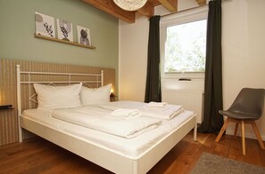 Un lit bien fait, avec du linge de lit blanc et des oreillers, une tête de lit en bois, une table de chevet avec une lampe, une chaise et une fenêtre avec des rideaux.