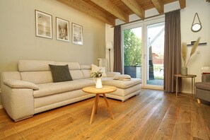 Ein Wohnzimmer mit einer Couch, einem kleinen runden Tisch und einer Blumenvase. An der Wand hängen drei gerahmte Bilder, ein Fenster mit Vorhängen und eine Holzbalkendecke.