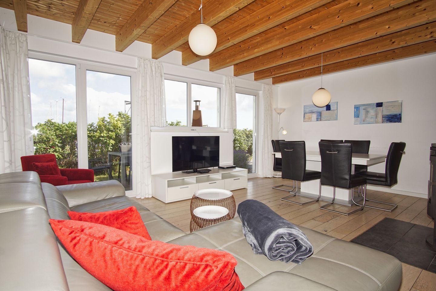 Un salon moderne doté d’un grand téléviseur, d’un canapé, de coussins rouges, d’un coin repas avec des chaises et de grandes fenêtres offrant une vue sur la verdure.