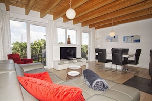 A modern living room with a large TV, a sofa, red cushions, a dining area with chairs, and large windows offering a view of greenery.