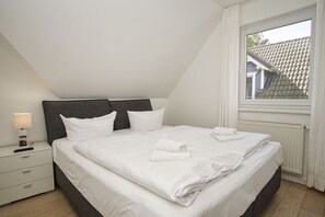 A neatly made bed in a room with a sloped ceiling, a nightstand with a lamp, and a window with a view of a house. - Vacation home near the harbor with terrace and sea view - Ferienhaus TimpeTe Backbord - (Gager)