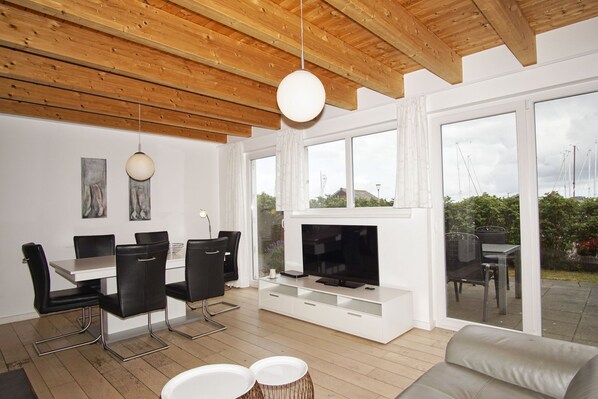 A modern living room with a wooden ceiling, a fireplace, a dining area with black chairs, and large windows offering a view of the outdoors.