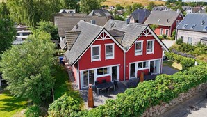 Une maison rouge avec un toit en pente, un porche à l’avant et un jardin aménagé avec une table et des chaises.