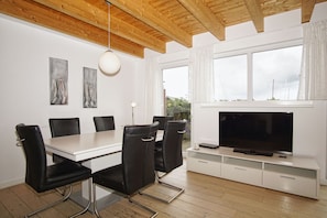 Ein moderner Essbereich mit einem weißen Tisch und schwarzen Stühlen, einem Flachbildfernseher auf einem weißen Schrank und Holzbalkendecken.