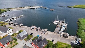 Vue aérienne d’une marina avec des bateaux amarrés, une jetée s’avançant dans l’eau et des bâtiments le long du littoral.
