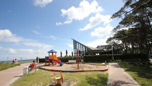 A playground with a slide and climbing structures, surrounded by a paved path and greenery.