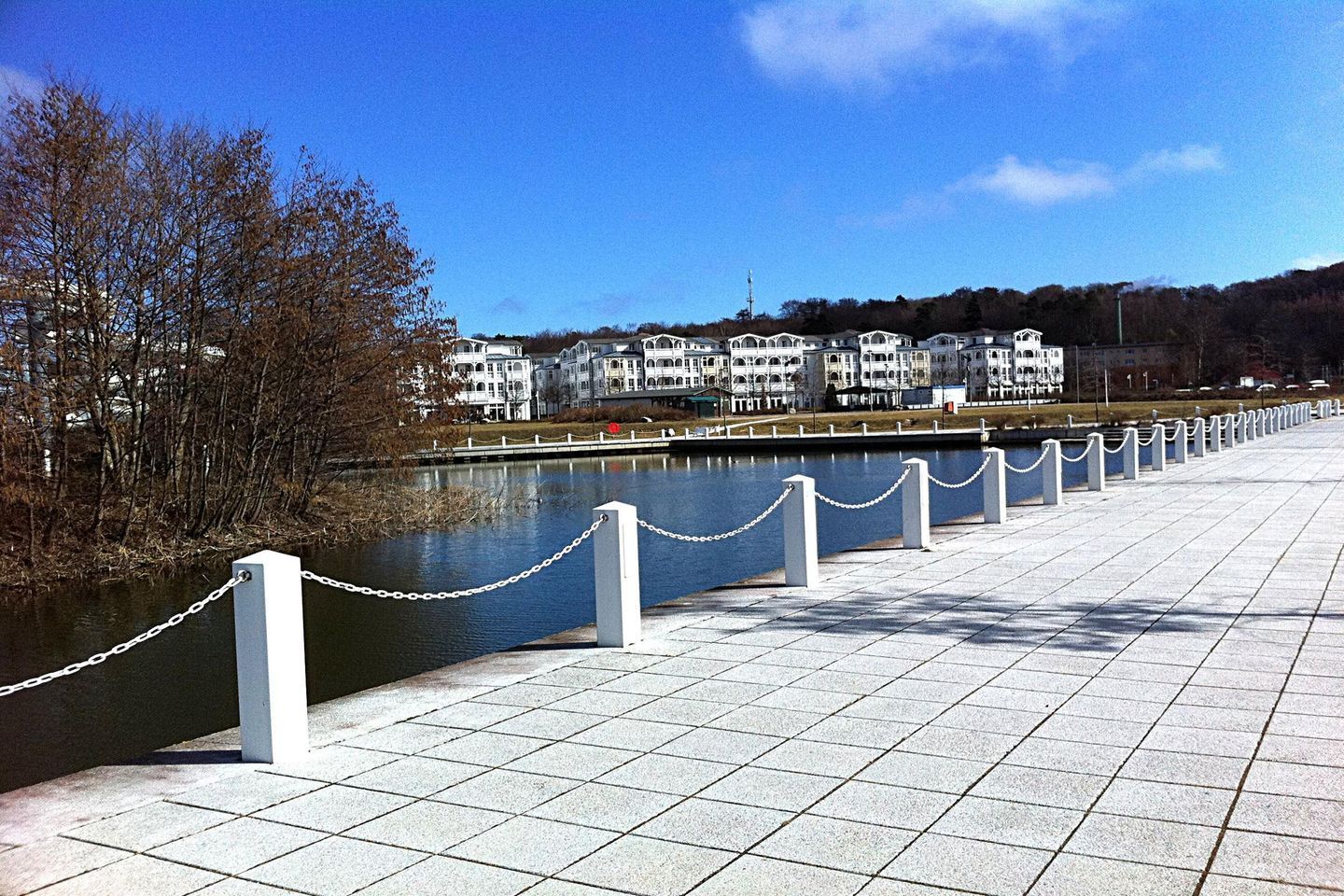 A waterfront promenade with a chain barrier, a river, and a row of white buildings in the background.