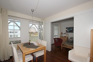 A spacious living room with a wooden dining table, white chairs, a red armchair, and a glass coffee table. A large window offers a view of the outside. - F-1044 Ferienwohnung Having - Ferienwohnung Having F-1044 Wohnung 315 (Sellin)