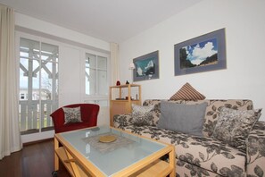 A living room with a glass coffee table, a red armchair, and a floral sofa. There are two framed pictures on the wall and a wooden side table with a lamp. - F-1044 Ferienwohnung Having - Ferienwohnung Having F-1044 Wohnung 315 (Sellin)
