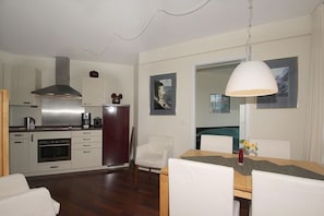 A modern kitchen with stainless steel appliances, a dining area with a wooden table and white chairs, and a large window with a view of the city. - F-1044 Ferienwohnung Having - Ferienwohnung Having F-1044 Wohnung 315 (Sellin)
