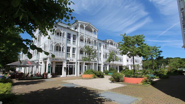 A large, white, multi-story building with a central courtyard, outdoor seating area, and a pizza restaurant.