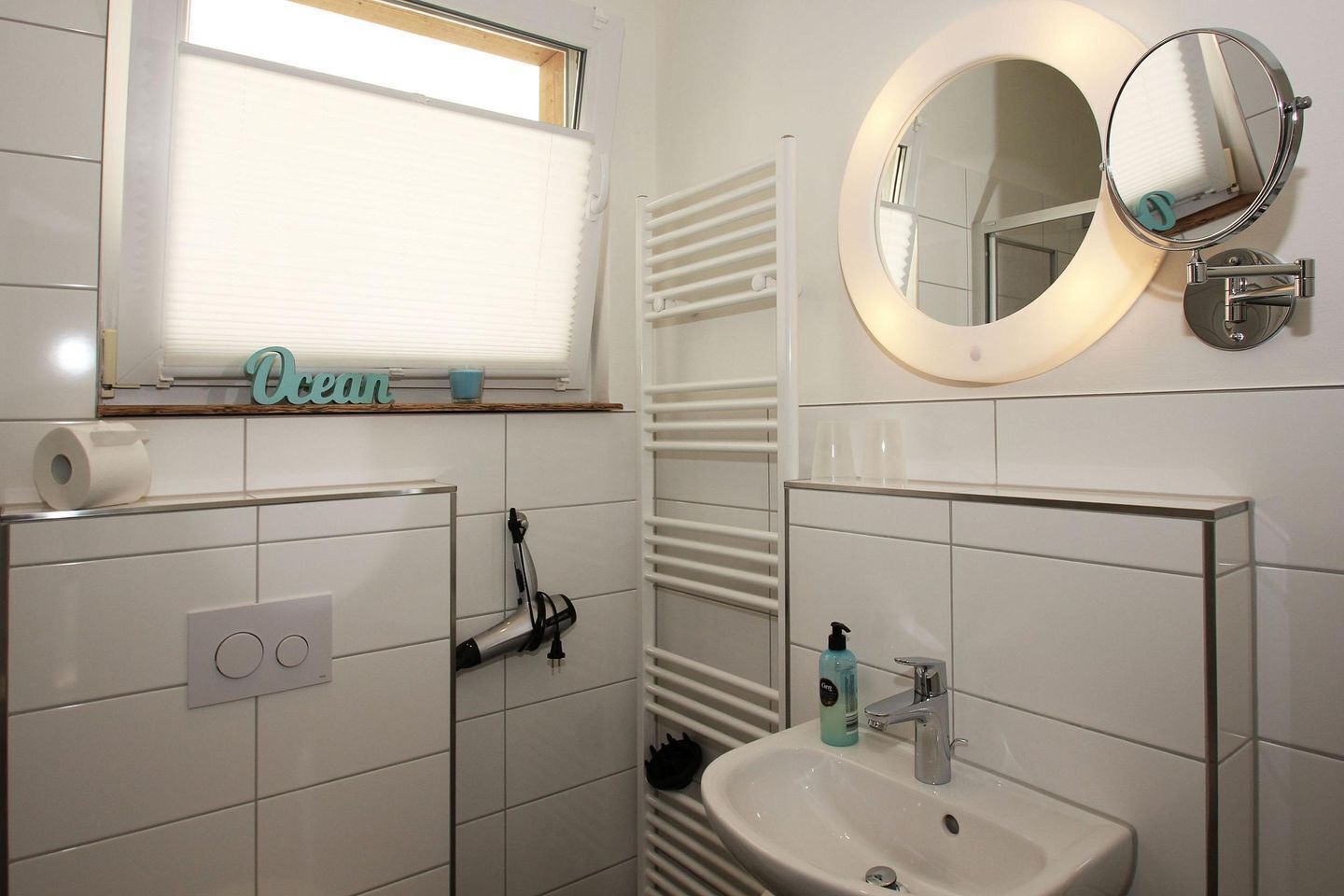 A bathroom with a toilet paper holder, a towel rack, a sink with a mirror, and a window with blinds.