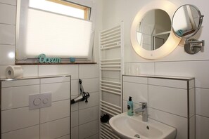A bathroom with a toilet paper holder, a towel rack, a sink with a mirror, and a window with blinds.