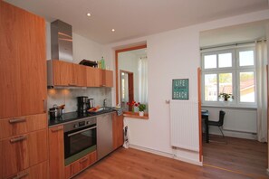 A modern kitchen with wooden cabinets, a built-in oven, and a coffee maker. There is a window with a view of buildings and a sign on the wall that says "Life is good at the beach".